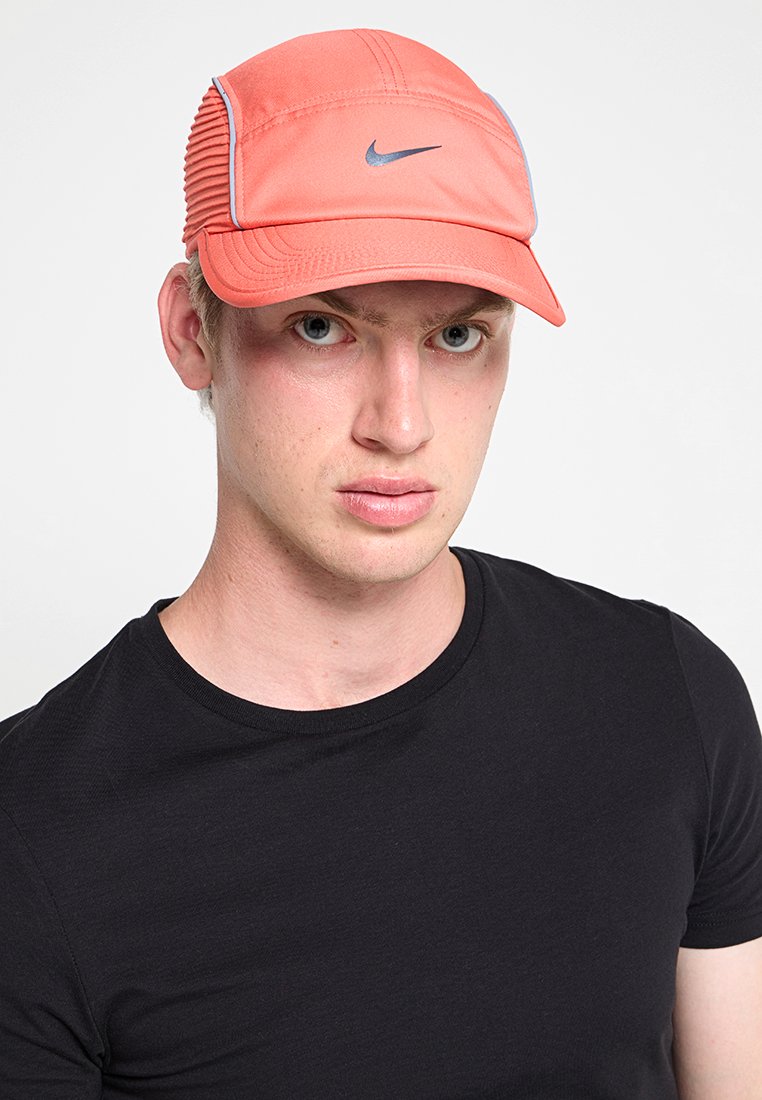 Jeune homme portant une casquette Nike corail et un t-shirt noir, faisant face à l'avant avec une expression neutre sur un fond clair.
