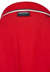 Rotes Polohemd mit flachem Kragen und einer weißen sowie schwarzen Akzentbordüre. Das Etikett "HECHTER PARIS" ist nah am Kragen aufgestickt.
