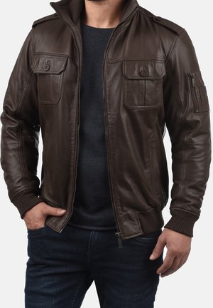 Homme portant une veste en cuir marron foncé avec poches poitrine boutonnées, poche à fermeture éclair sur le poignet, col côtelé, et jean foncé.