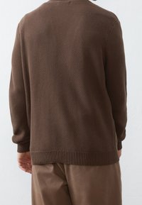 Brauner gestrickter Pullover mit strukturiertem Muster, gerippten Bündchen und Saum. Die Rückseite zeigt eine lockere Passform, die über beigen Hosen getragen wird.