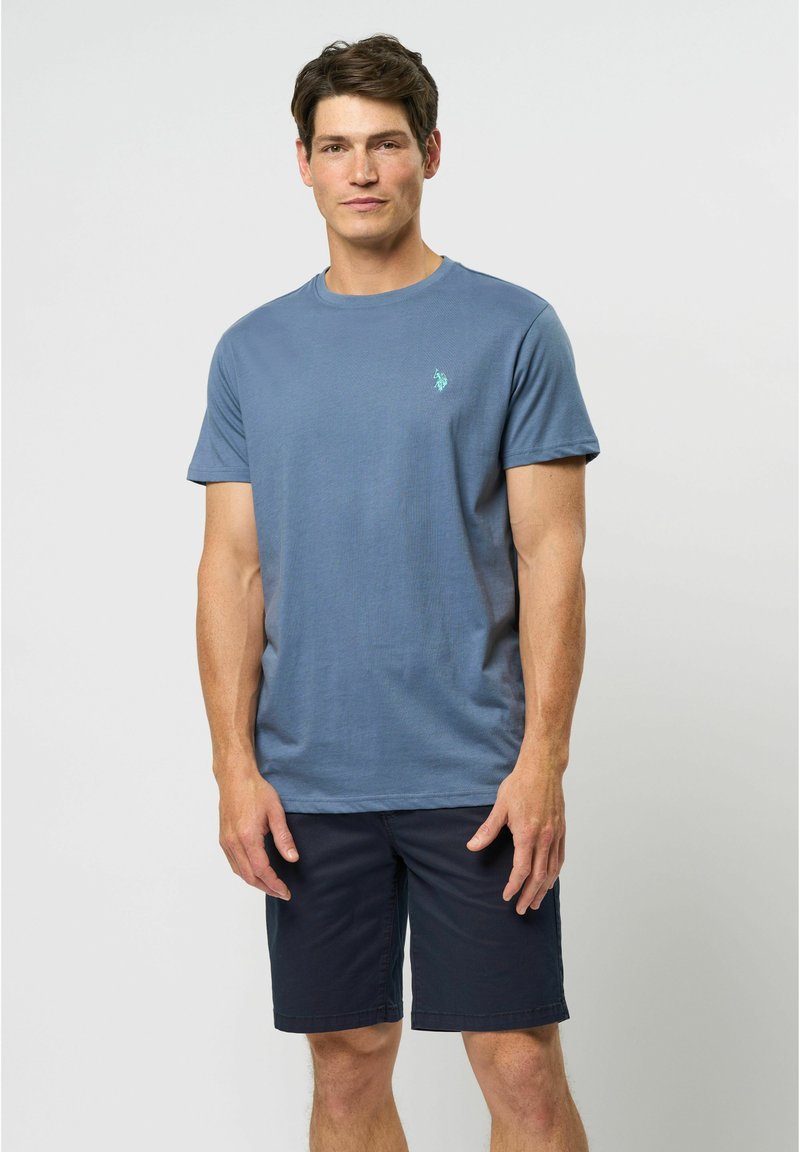 Pánské tričko s kulatým výstřihem v jasně modré bavlně, s malým zeleným logem na hrudi. Spárováno s tmavě námořnickými šortkami.