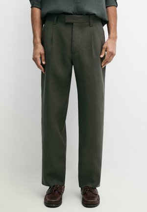 Uomo che indossa pantaloni dritti verde oliva e scarpe da barca in pelle marrone, in piedi davanti a uno sfondo bianco semplice.