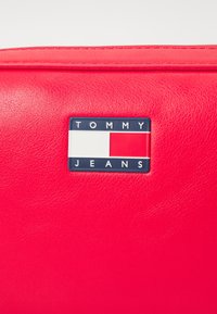 Sac en cuir rouge avec un logo bleu rectangulaire portant le texte "TOMMY JEANS" et un design en blocs de couleurs blanc et rouge. Texture lisse.