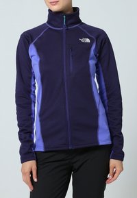 Fioletowa polarowa kurtka z wysokim kołnierzem, pełnym zamkiem błyskawicznym i kontrastującymi, jaśniejszymi fioletowymi panelami bocznymi. Posiada kieszeń na piersi i teksturowany materiał.