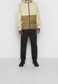 Dwukolorowa kurtka w beżowo-oliwkowym odcieniu z kapturem, przednim zamkiem błyskawicznym, elastycznymi mankietami i nadrukowanym logo; w zestawieniu z ciemnymi spodniami i czarnymi butami.