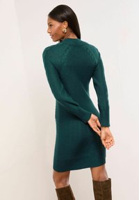 Tirkīzzils adīts kleita ar ribotām apakšmalām un piedurknēm, kura piedāvā pieguļošu siluetu un kabelīšu adījumus plecu un muguras daļā.