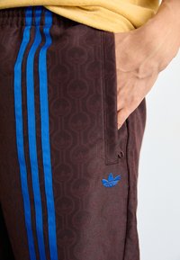 Rudos raštuoti sportiniai kelnes su mėlynomis juostelėmis šonuose ir mėlynu logotipo elementu. Turi šoninį kišenę ir elastingą juosmenį.