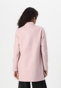 Cappotto lungo rosa chiaro con colletto alto e maniche lunghe. Realizzato in un tessuto morbido e texture. Design semplice, senza chiusure o motivi visibili.