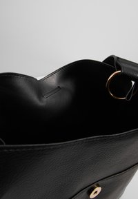 Czarna torba typu tote ze skóry o gładkiej fakturze, z zaokrąglonym górnym brzegiem i złotym, okrągłym elementem ozdobnym przy uchwycie.