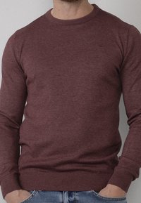 Uomo che indossa un maglione girocollo bordeaux aderente, con polsini e orlo a coste, mani infilate nelle tasche di jeans azzurri chiari.