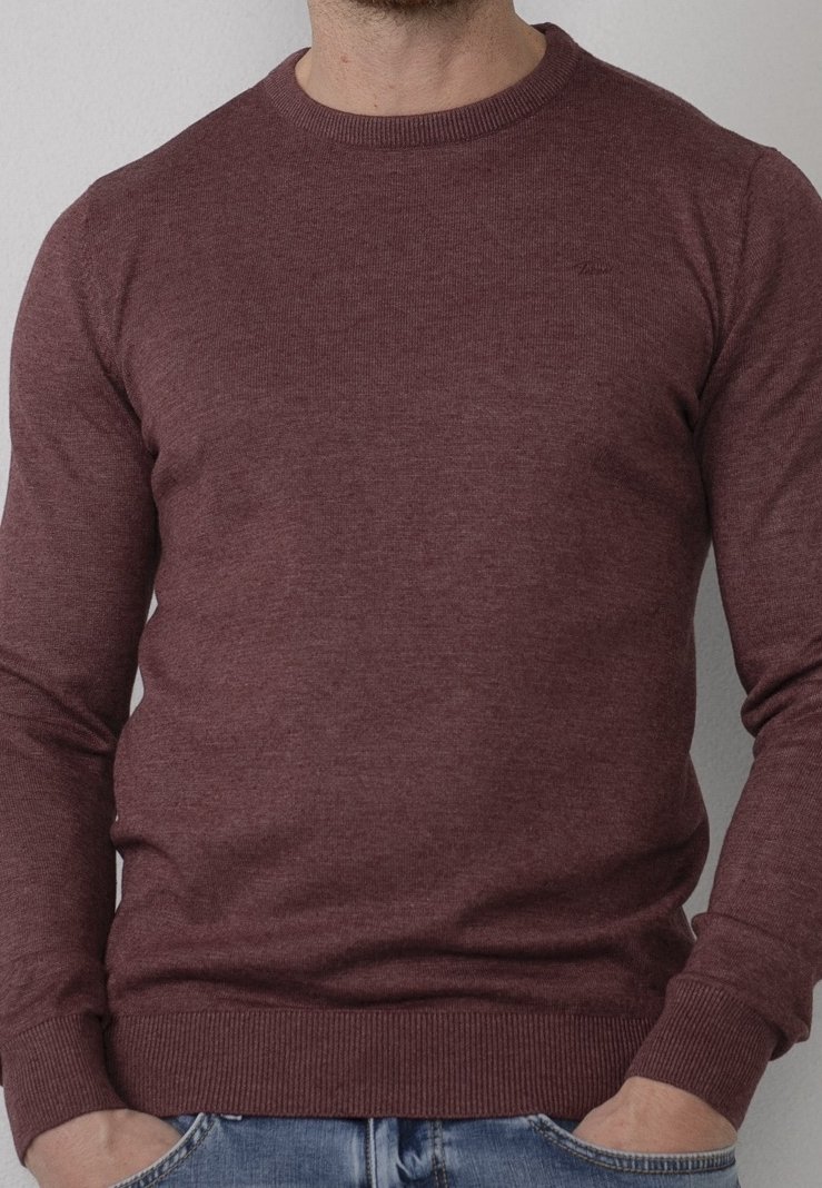 Uomo che indossa un maglione girocollo bordeaux aderente, con polsini e orlo a coste, mani infilate nelle tasche di jeans azzurri chiari.