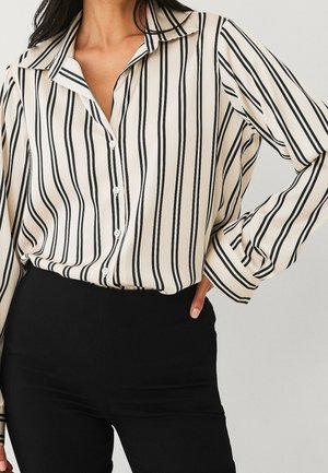 Femme portant un chemisier crème à rayures verticales noires rentré dans un pantalon noir taille haute, une main sur la hanche.