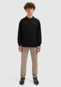 Zwarte polo met lange mouwen en kraag, met logo op de linker borst; gecombineerd met beige strakke broek en zwarte sportschoenen.