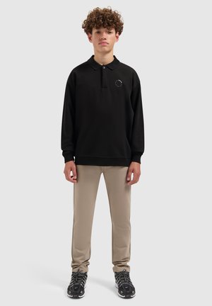 Zwarte polo met lange mouwen en kraag, met logo op de linker borst; gecombineerd met beige strakke broek en zwarte sportschoenen.