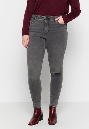 Personne portant un jean gris ajusté, un haut bordeaux à manches longues et des bottines bordeaux brillantes, debout avec une main dans la poche.