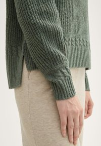 Zielony sweter w dzianinie z prążkowanymi mankietami i bocznymi rozcięciami, o fakturowanym wzorze i luźnym kroju, w zestawieniu z beżowymi spodniami.