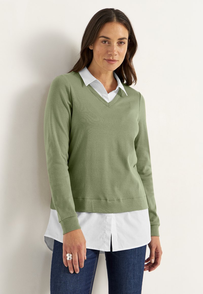Lichtgroene v-hals trui van zachte stof, gedragen over een wit overhemden, in combinatie met donkere jeans. Heeft lange mouwen en geribde manchetten.