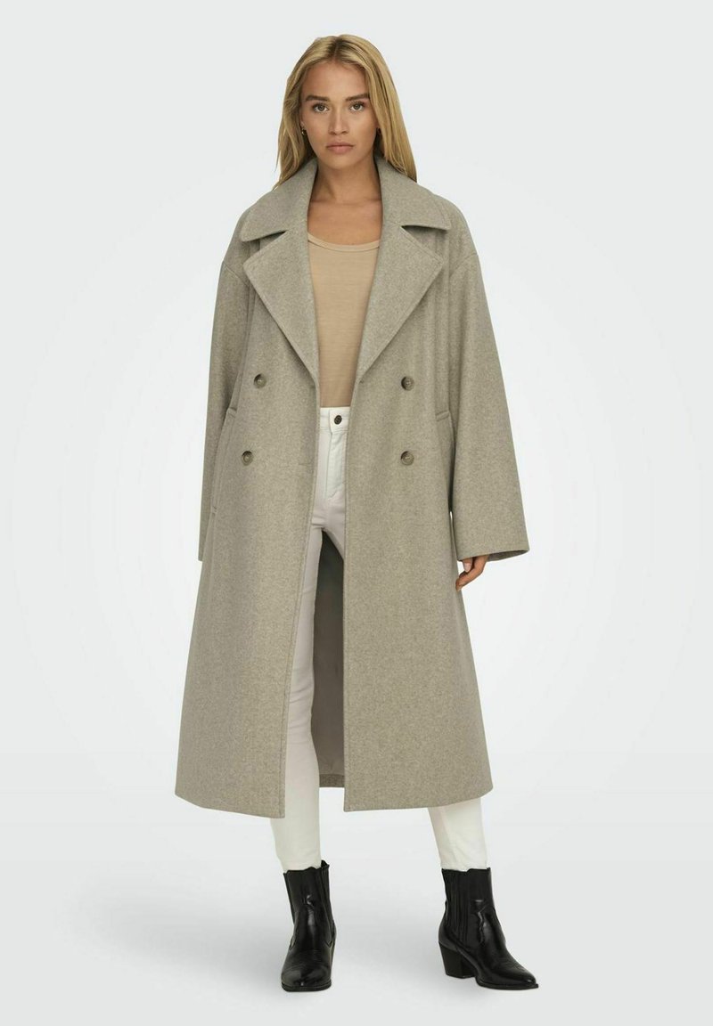 Lange grijze wollen jas met een dubbelknoopdesign, brede revers en zijzakken, gedragen over een beige top en witte broek. Zwarte enkellaarsjes.