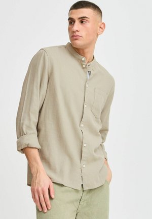 Chemise boutonnée beige clair avec un col mandarin, manches longues retroussées aux poignets et une poche poitrine. Tissu doux et texturé.