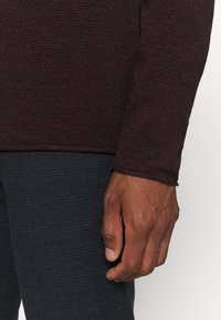 Maglione bordeaux a trama con maniche lunghe, esposto accanto a pantaloni scuri con motivo. Dettaglio di una mano appoggiata sui pantaloni.