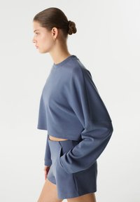 Zkrácená modrá mikina s dlouhými rukávy, měkkou texturou a minimálními švy. Vybavená odpovídajícími modrými kraťasy, oba kousky mají ležérní design.