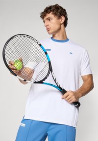 Hvid tennis skjorte med blå krave, parret med lyseblå shorts. Personen holder et sort tennisracket og en grøn tennisbold.