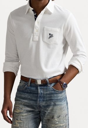 Weißes Langarm-Polohemd mit einer Tasche und einem dunkelblauen Logo, kombiniert mit zerrissenen blauen Jeans und einem braunen Gürtel. Uhr am Handgelenk sichtbar.