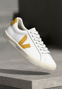 Valged sneakers, mis on valmistatud siledast materjalist, külgedel kollase kaldu logoga, tekstureeritud kummist tallaga ja lukkudega, et tagada kindel istuvus.