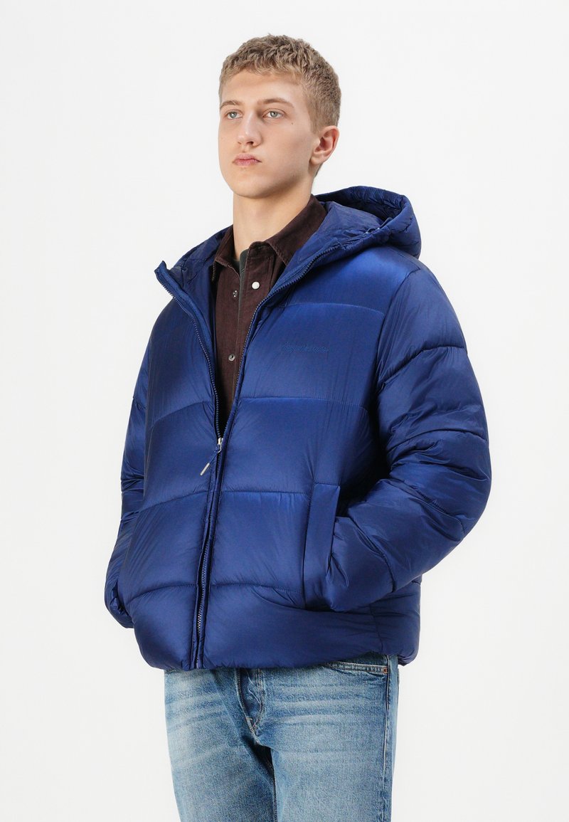 Blauer Puffer-Jacke mit Kapuze, mit gestepptem Design und Frontreißverschluss, getragen über einem dunklen Hemd, kombiniert mit hellblauen Jeans.