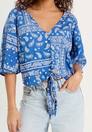 Femme portant un chemisier bleu à motifs boutonné, noué à la taille, avec un jean clair, main dans la poche et cheveux bouclés visibles aux épaules.