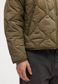 Brazo izquierdo y mano de una persona que lleva una chaqueta acolchada verde oliva y jeans de mezclilla gris oscurecido, contra un fondo liso.