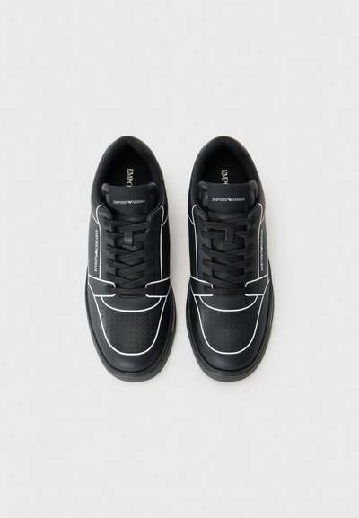 Baskets en cuir noir avec des accents blancs, présentant un bout perforé, des lacets ronds et un logo sur la languette. Texture lisse et design sportif.