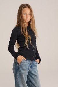 Juodas raukinis kardiganas su mygtukų uždarymu, su banguotais rankogaliais, dėvimas virš laisvų mėlynų džinsų. Modelis pozuoja su rankomis kišenėse.