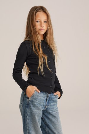 Zwarte geribbelde cardigan met knoopsluiting en gegolfde manchetten, gedragen over een blauwe losse denim jeans, model poseert met handen in de zakken.