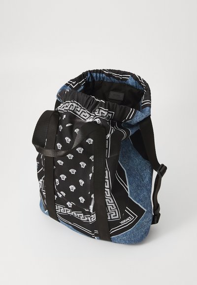 Zwarte en blauwe rugzak met trekkoord bovenaan, denimtextuur, gemengde patronen waaronder een bandana-print, en zwarte schouderbanden.