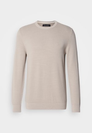 Jersey de punto fino en color beige de manga larga y cuello redondo, con un logo sutil en el pecho, presentado sobre un fondo blanco liso.