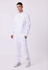 Witte sweatshirt en sweatpants met lichtblauwe stiksels. De sweatshirt heeft een ronde hals en een subtiele logo. Schoenen zijn lichtgrijs met een gestructureerd ontwerp.