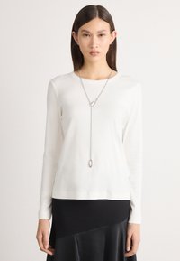 Langärmliger, weißer Pullover aus weichem Material mit rundem Ausschnitt. Eine Silberkette mit ovalem Anhänger hängt auffällig.