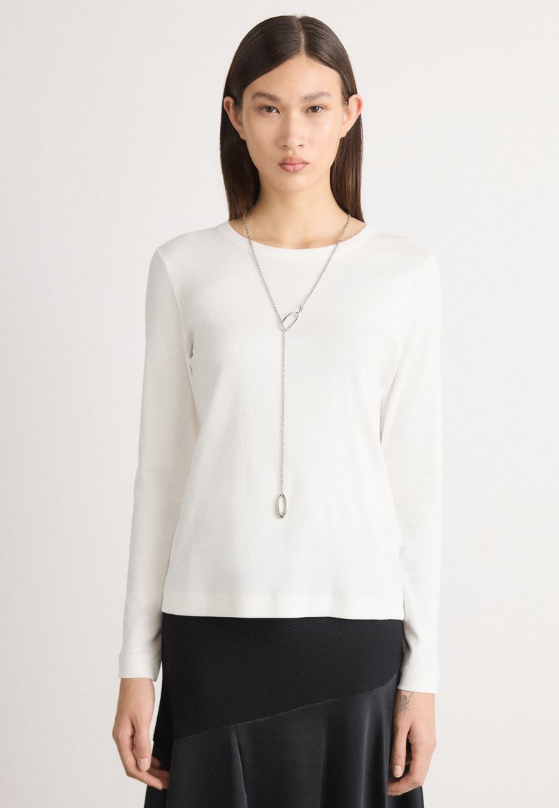 Langärmliger, weißer Pullover aus weichem Material mit rundem Ausschnitt. Eine Silberkette mit ovalem Anhänger hängt auffällig.