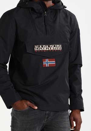 Mężczyzna noszący czarną kurtkę anorak marki Napapijri z klapką na przedniej kieszeni i naszywką norweskiej flagi, z rękami w kieszeniach ciemnych dżinsów.