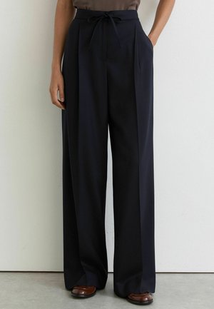 Schwarze, hoch taillierte Hose mit weitem Bein, vorne mit Falten und Tunnelzugbund, kombiniert mit braunen Lederschuhen und einem taupenfarbenen Oberteil.