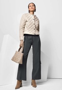 Beige gesteppte Jacke mit Rautenmuster, graue weit geschnittene Hose, beige Handtasche und hellbraune Absatzstiefel vor einem minimalistischen Hintergrund.