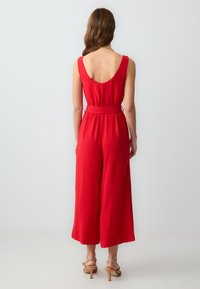 Roter Jumpsuit mit Rundrücken, ärmellosem Design und tailliertem Bund. Verfügt über weite Hosenbeine und ein leichtes Material. Rückansicht gezeigt.