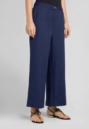 Persona che indossa pantaloni a gamba larga blu navy con vita elastica e chiusura con bottoni, abbinati a sandali neri piatti decorati con perline.