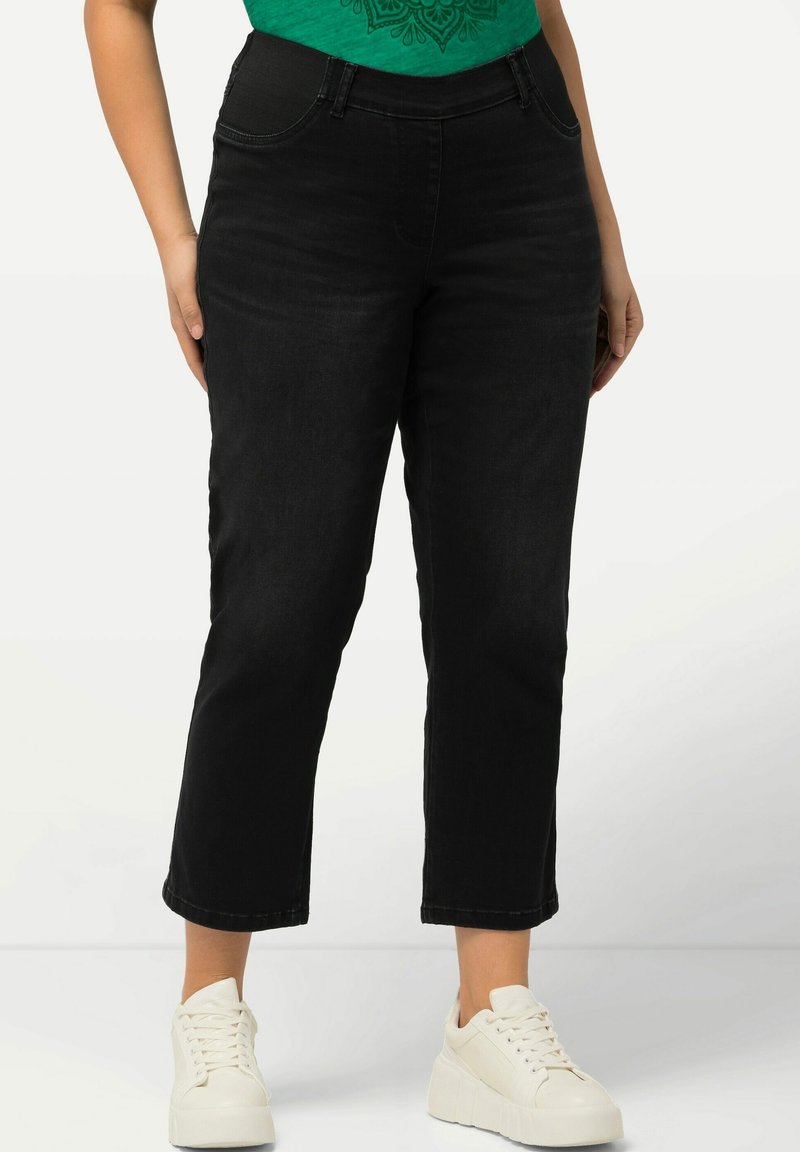 Pantalones de mezclilla negros con cintura ajustada y piernas rectas ligeramente recortadas. Combinados con una camiseta gráfica verde y zapatillas blancas.