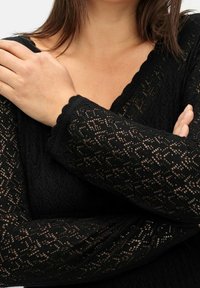 Černý pletený top z krajky s výstřihem do V, složitými vzory a vroubkovanými lemy rukávů, představující lehkou a texturovanou látku.