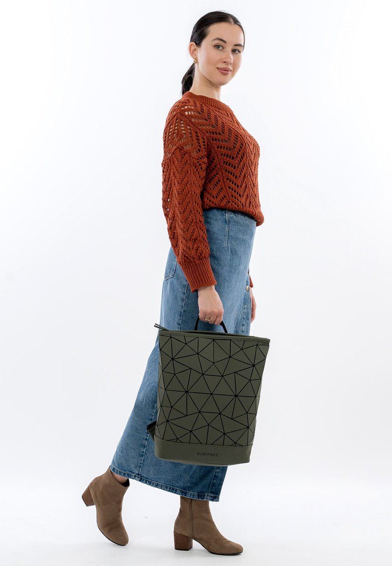 Bolso tote verde oliva con patrón geométrico negro, material de goma y asa superior. Llevado por una modelo con suéter de punto naranja y falda vaquera.