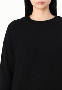 Eine Frau trägt einen weichen schwarzen Rundhalsausschnitt-Pullover, gezeigt von den Schultern bis zu den Lippen, vor einem schlichten weißen Hintergrund.
