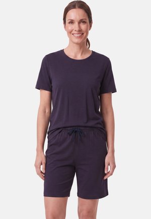 Lachende vrouw met bruin haar in een staart draagt een donkerpaarse korte mouwen shirt en bijpassende short met trekkoord tegen een effen achtergrond.