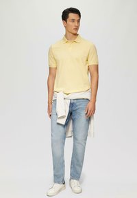 Polo shirt jaune à manches courtes, en coton, avec un col ; associé à un jean en denim bleu clair et des baskets blanches, avec un léger pull noué à la taille.
